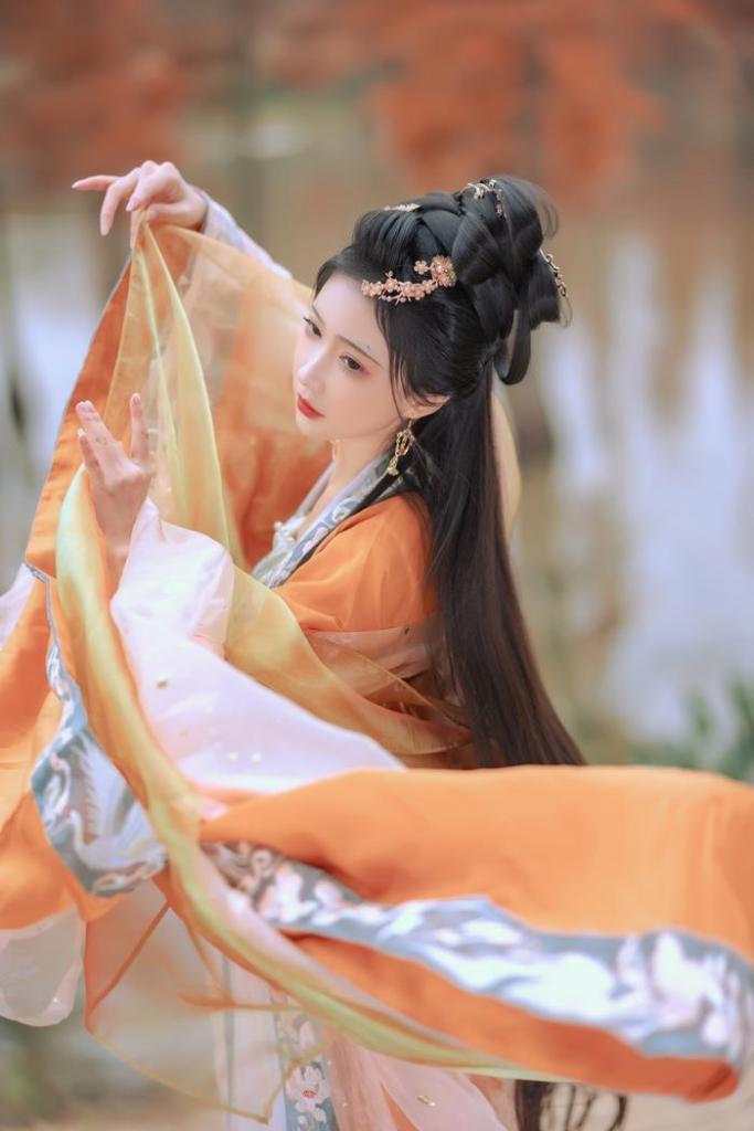 A woman in traditional attire gracefully dancing, wearing an orange and flowy garment, with her long dark hair styled elegantly, set against a serene natural backdrop.