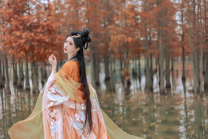 A woman in traditional orange and white attire standing near a body of water, surrounded by autumn trees with orange leaves.