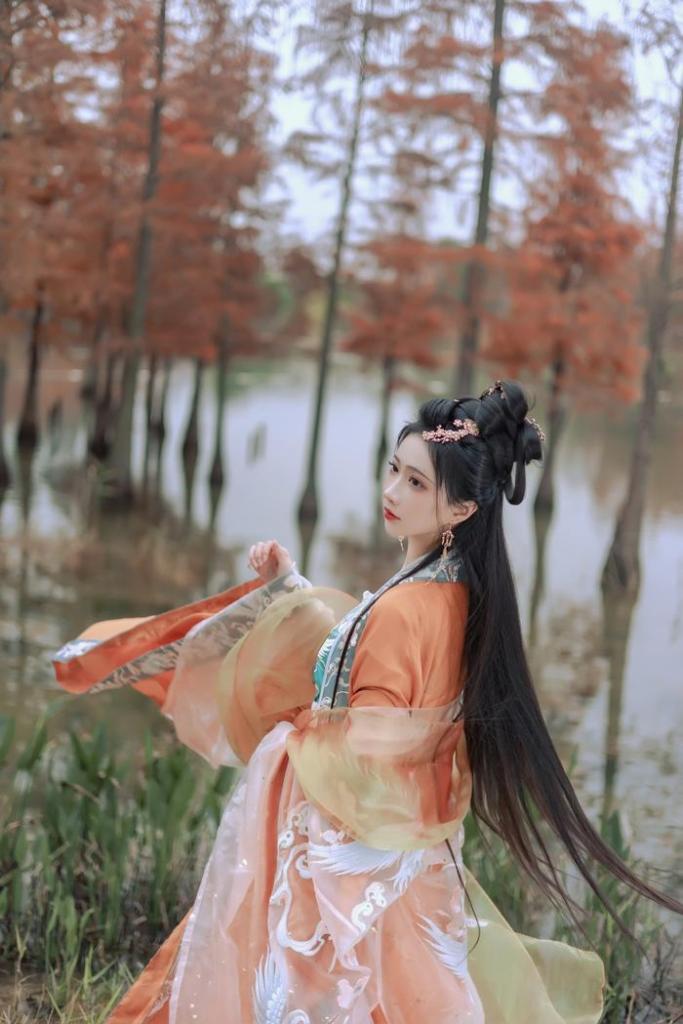 A woman in traditional attire gracefully poses by a serene body of water surrounded by autumn foliage.