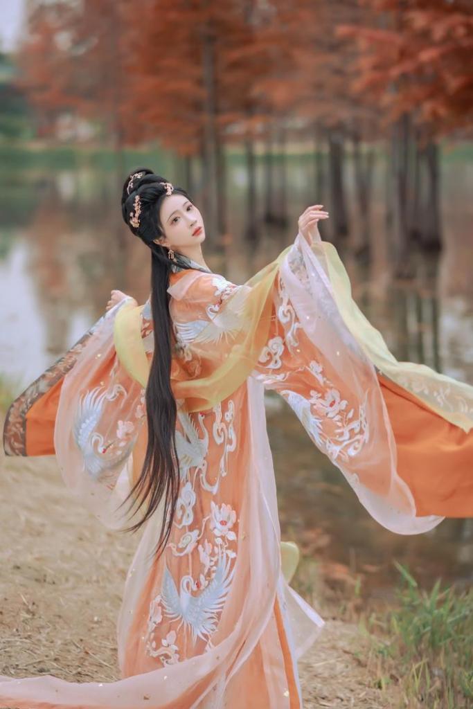 A woman in traditional attire with flowing sleeves stands gracefully by a serene pond surrounded by autumn foliage.