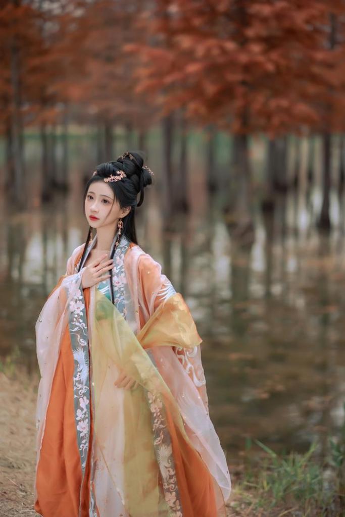 A young woman in a traditional, colorful outfit stands by a serene body of water, surrounded by trees with orange leaves, showcasing a peaceful and picturesque autumn setting.