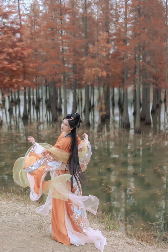 A woman in traditional dress, with long hair, dances gracefully near a calm body of water surrounded by autumn trees.