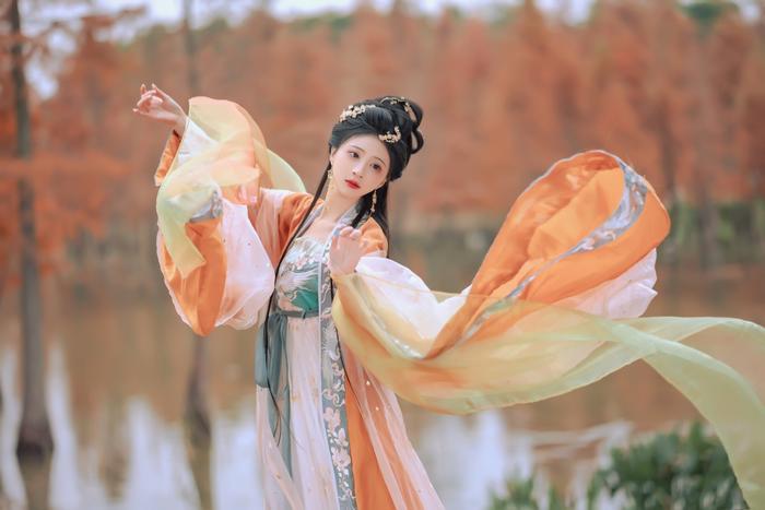 A woman dressed in a traditional Chinese outfit with flowing sleeves dances gracefully near a serene water body, surrounded by autumn-colored trees.