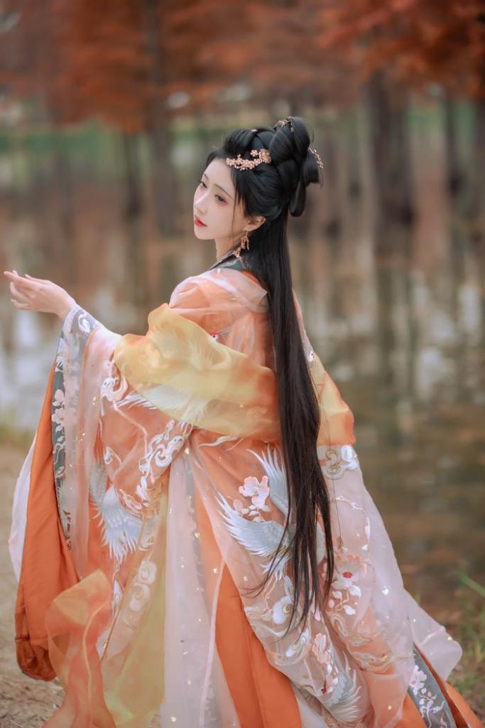 A woman wearing a traditional orange and cream colored hanfu stands by a body of water, with long hair and intricate accessories, set against a backdrop of autumn foliage.