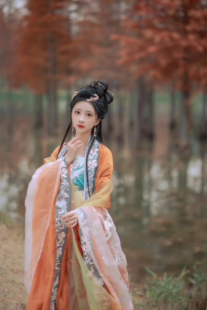 A woman in traditional Asian attire stands by a serene body of water, surrounded by autumn trees with orange leaves.
