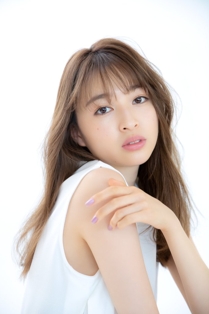 A woman with long, wavy hair, gazing directly at the camera with a soft expression. She is wearing a white sleeveless top and has her hand softly placed on her shoulder.