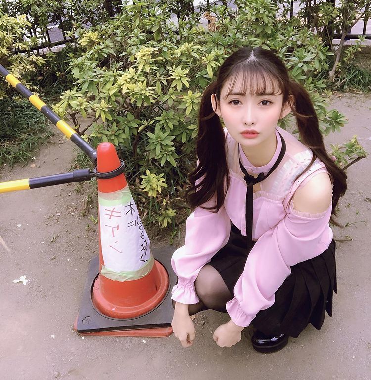 A young woman with long hair styled in pigtails is crouching next to a traffic cone surrounded by greenery. She is wearing a pink blouse with a black skirt and has a serious expression on her face.