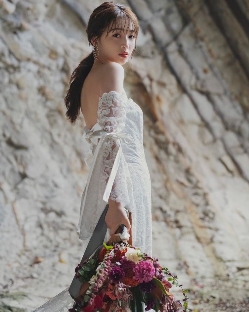 A woman in a white off-shoulder dress poses against a rocky background, holding a vibrant bouquet of flowers.