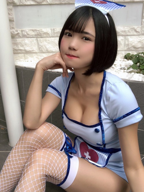 A young woman dressed in a nurse costume, sitting with her chin resting on her hand, in a relaxed pose. The outfit is light blue with a white and blue headpiece, and she is accessorized with fishnet stockings.