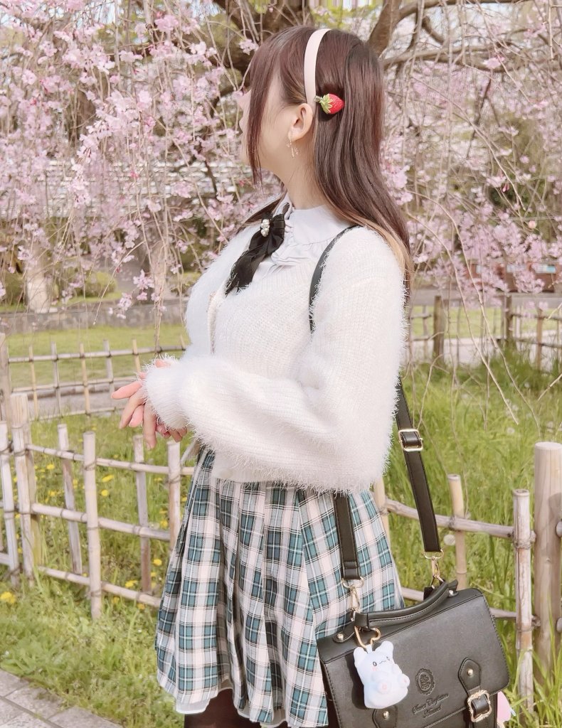A person standing in front of blooming cherry blossom trees, wearing a fluffy white sweater, plaid skirt, and a bag with a cute charm.