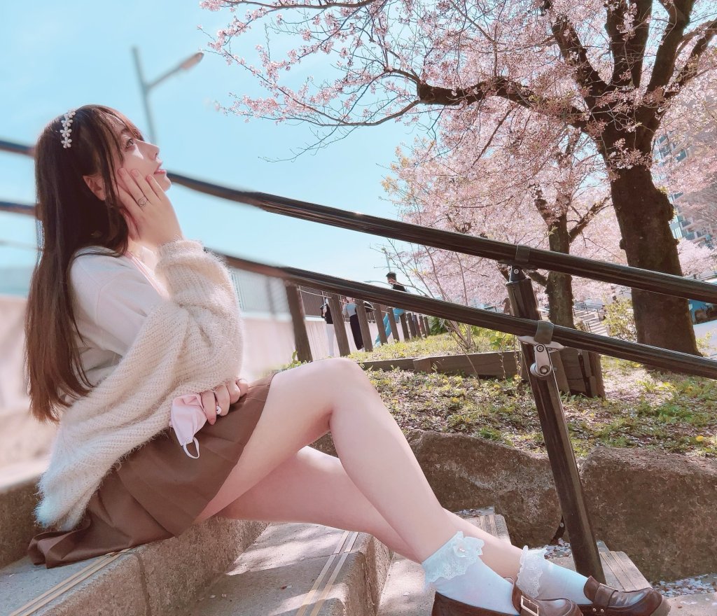 A young woman sitting on steps in a park, wearing a cozy sweater and a skirt, looking thoughtfully at cherry blossom trees in full bloom.