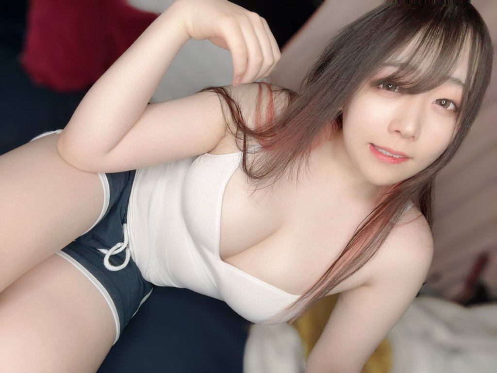 A person with long hair styled down, wearing a white tank top and black shorts, posed while sitting on a bed with soft bedding.