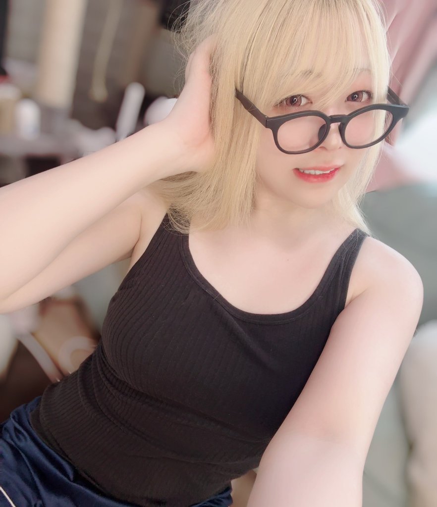 A person with blonde hair wearing black glasses and a black tank top, smiling and posing for the camera.