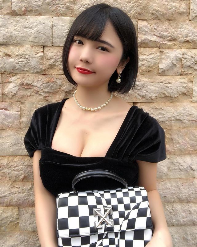 A woman with short black hair wearing a black dress and a pearl necklace, holding a black and white checkered handbag, against a stone wall.