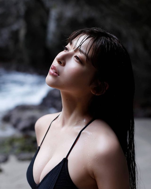 A woman with long dark hair gazes thoughtfully upwards, wearing a black swimsuit, with a rocky beach and ocean in the background.