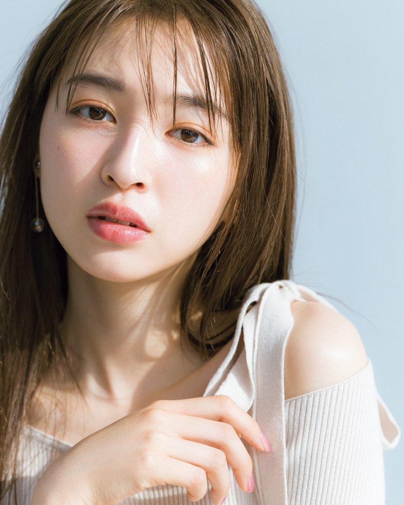 A close-up portrait of a woman with long brown hair and a natural makeup look, wearing a cream-colored top with shoulder straps. She is gazing directly at the camera with a soft expression.