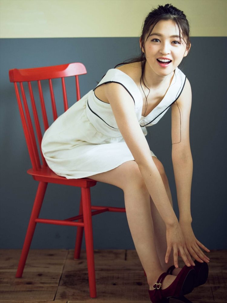 A young woman in a white dress is sitting on a red chair, smiling while reaching down to adjust her shoe.