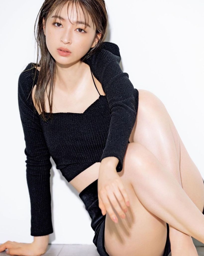 A young woman with long hair poses sitting on the floor, wearing a black cropped sweater and a matching skirt, against a plain white background.