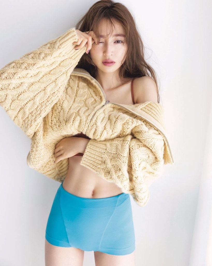 A person poses softly in a cozy oversized yellow knit sweater with a blue short bottom, showcasing a relaxed expression against a neutral background.