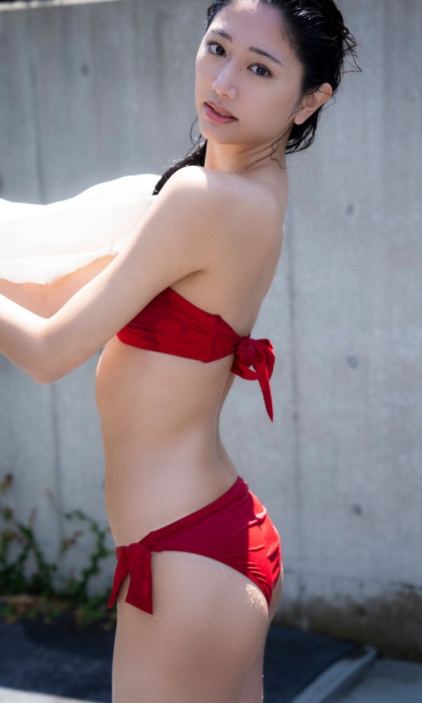 A young woman poses in profile wearing a red bikini set with bow details, standing outdoors with a concrete background.