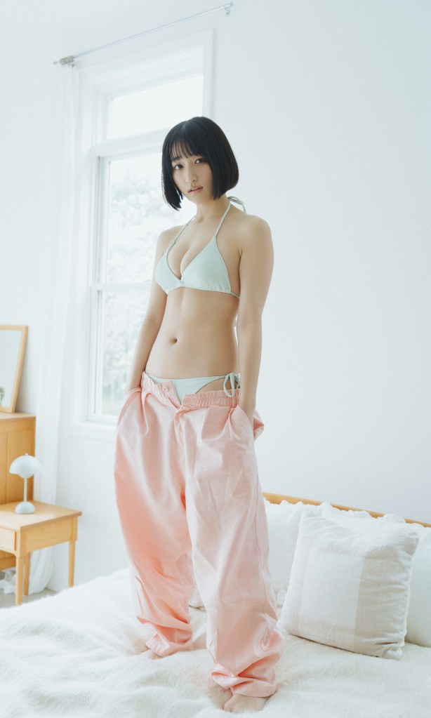 A young woman with a bob haircut wearing a light blue bikini top and oversized pink pants, standing on a bed in a bright room with large windows.