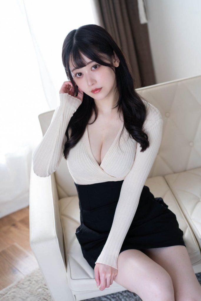 A young woman sitting on a sofa, wearing a fitted cream sweater and a black skirt, with her hand on her hair, and looking directly at the camera.