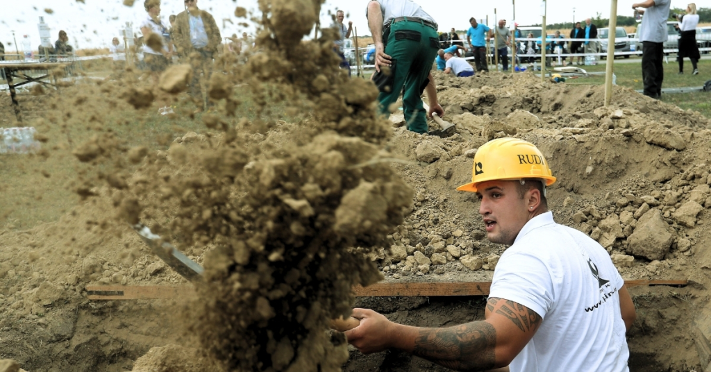 一名參賽者在挖墳大賽中挖掘土壤，周圍有其他參賽者和觀眾，並展示出比賽的激烈環境。