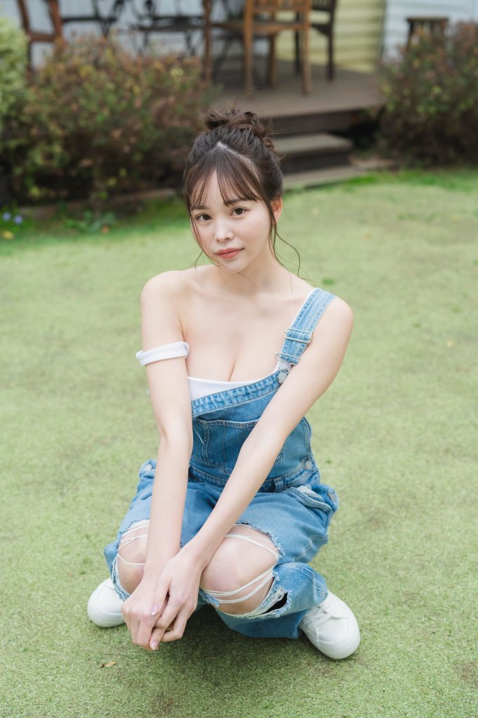 A young woman wearing a blue denim overall and white top is kneeling on lush green grass, looking at the camera with a soft expression.