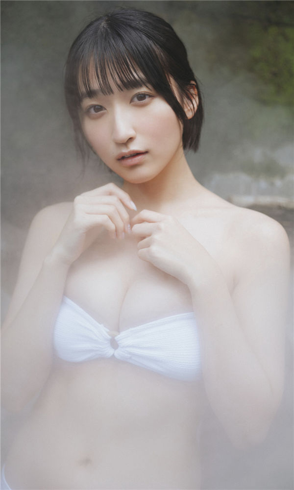 A young woman with shoulder-length black hair and bangs is posing in a misty setting, wearing a white bikini top. She has a gentle expression and is holding her hands close to her face.