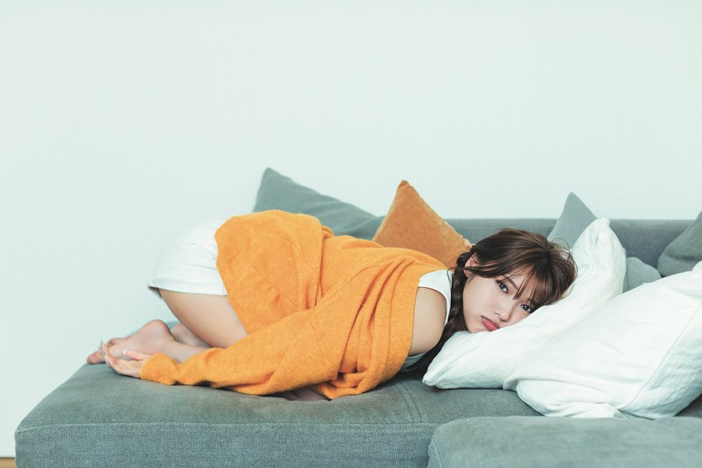 A person in an orange sweater and white shorts is lying on a gray couch, resting with their head on a pillow and looking directly at the camera.