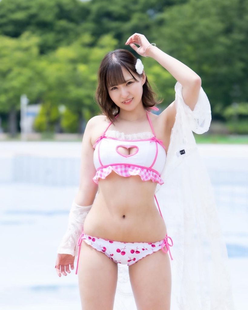 A woman poses in a playful bikini with a heart design and cherry patterns, standing outdoors with lush greenery in the background.
