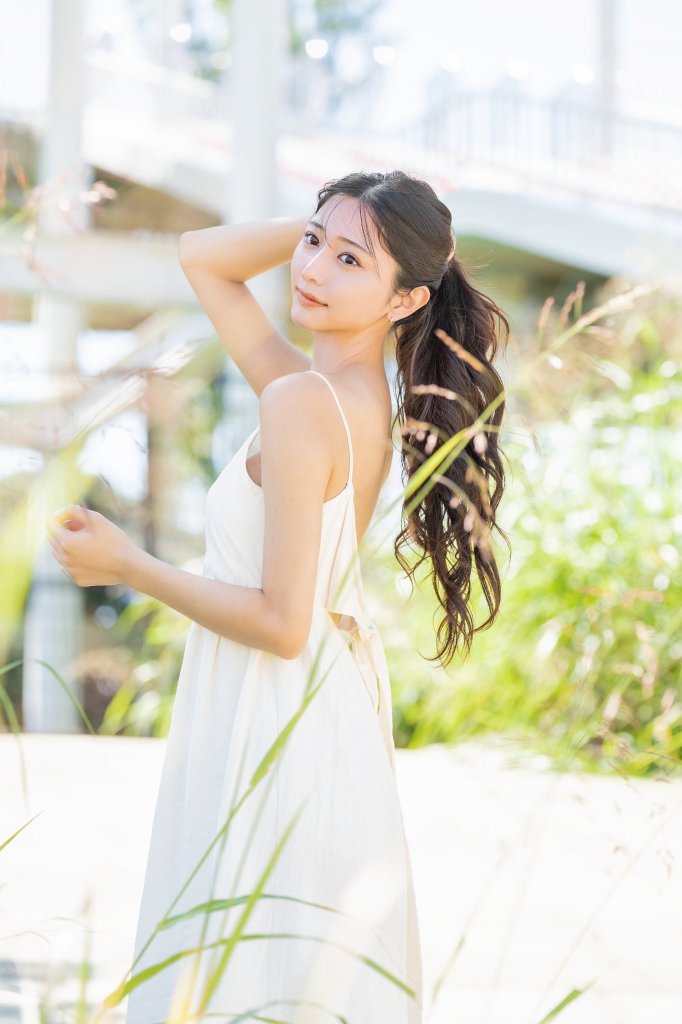 A young woman with long, dark hair stands outdoors, wearing a white dress. She has one hand behind her head and is looking towards the camera. Green grass and plants are in the foreground.