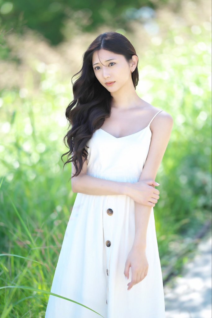 A young woman with long, wavy hair wearing a white dress stands in a green outdoor setting, with natural sunlight softly illuminating the scene.