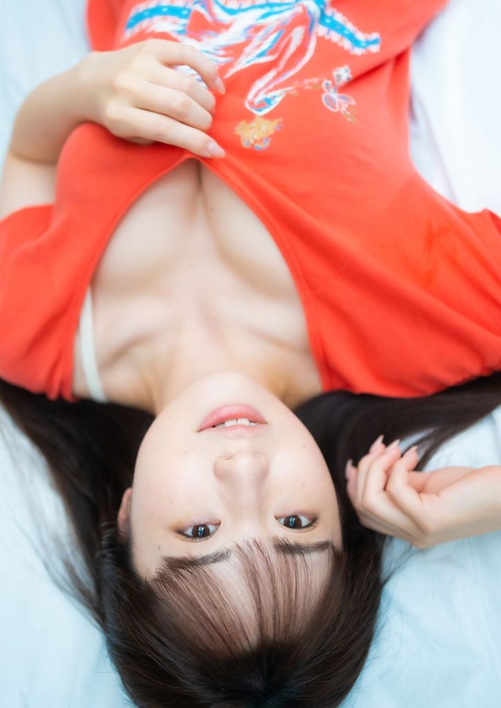 A person lying on a bed wearing a bright orange shirt with a design, appearing relaxed and looking up at the camera.