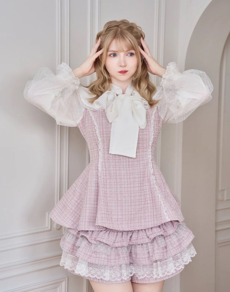 A young woman wearing a stylish pink peplum blouse with puffed sleeves and a large bow, accessorized with a frilled skirt. She is posing with her hands on her head in a softly lit, elegant indoor setting.