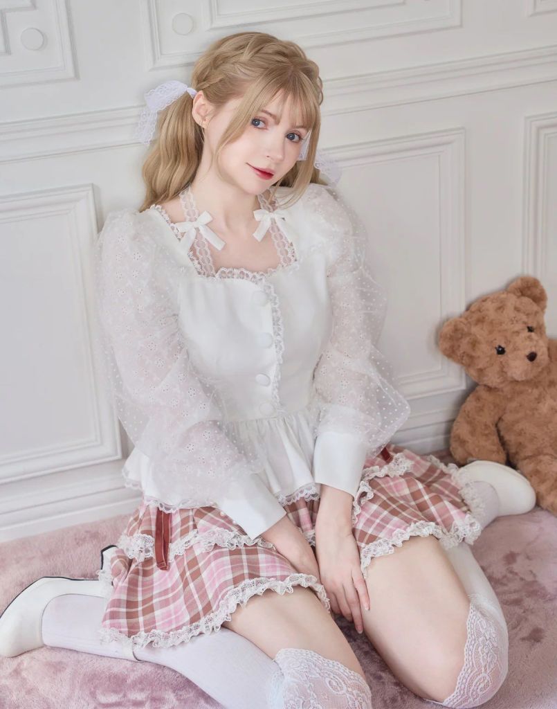 A young woman in a white blouse with puff sleeves and a pink plaid skirt, sitting on a soft surface beside a teddy bear, with her hair styled in loose waves and ribbons.