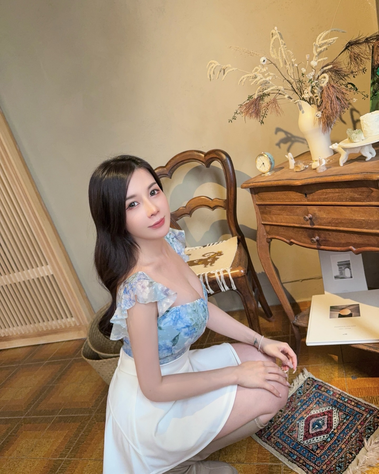 A woman wearing a floral blouse and white skirt is kneeling beside a wooden table decorated with items like a vase and a clock in a cozy indoor setting.