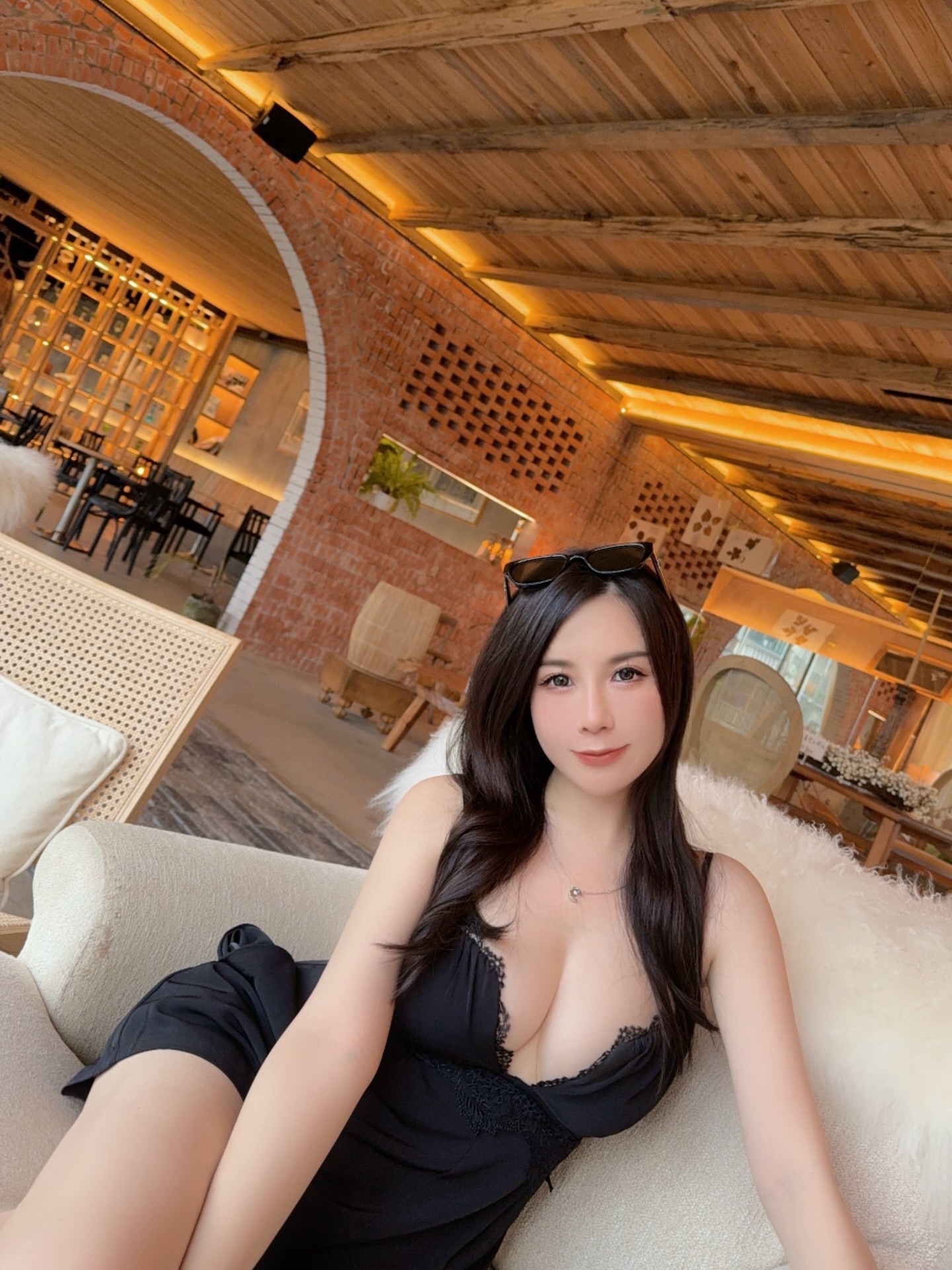 A woman in a black dress sitting on a couch in a stylish interior space with wooden beams and exposed brick walls.