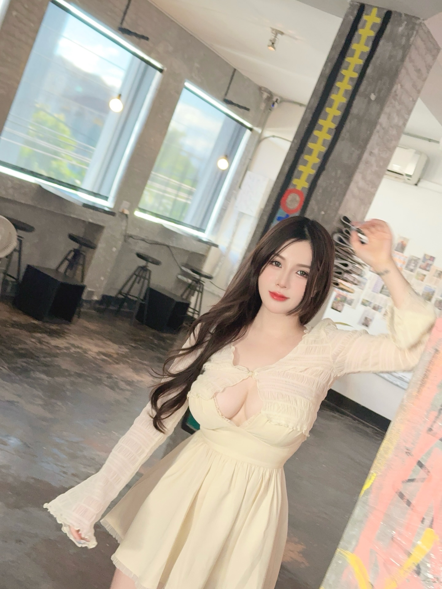 A young woman with long dark hair wearing a light yellow dress, posing in a modern interior with large windows and industrial decor.