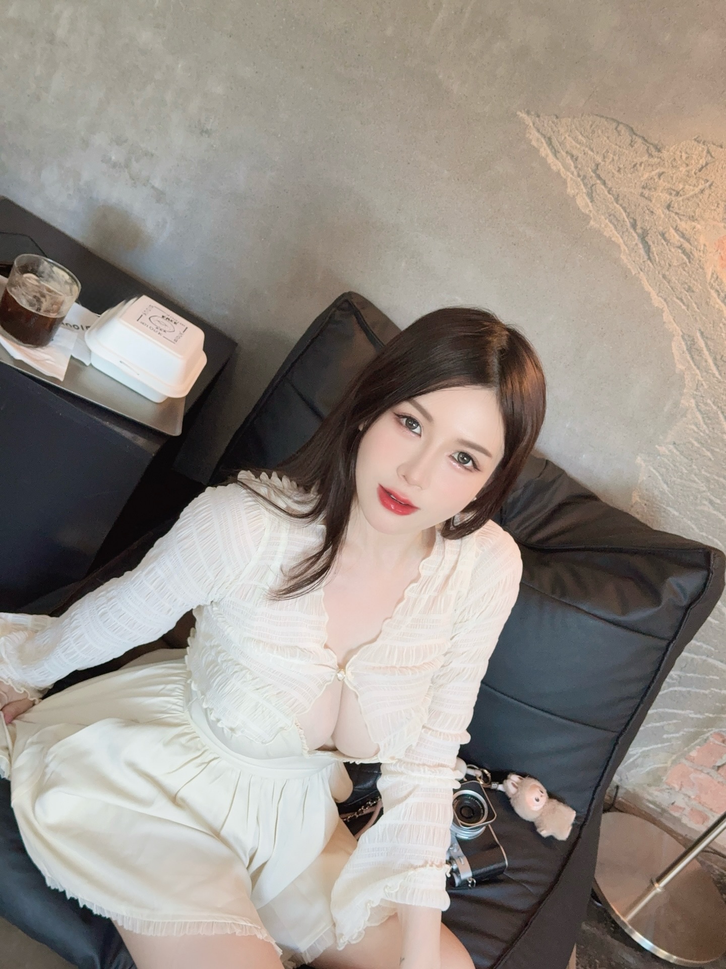 A woman wearing a white blouse and skirt sitting on a black sofa, looking directly at the camera, with a glass of drink and a camera beside her.