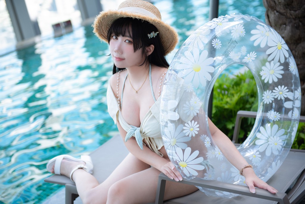 A woman in a swimsuit sits on a lounge chair by a pool, holding a clear inflatable ring decorated with daisies, wearing a straw hat.