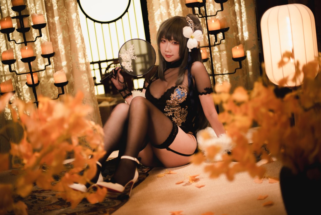 A person sitting on a decorated floor, wearing a black lace outfit with blue embellishments, holding a fan, surrounded by flowers and soft lighting.