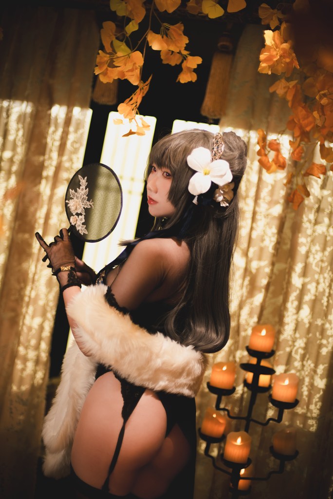 A woman in a stylish costume holding a fan, with long dark hair and a flower accessory, standing in front of a backdrop with sheer curtains and candles.