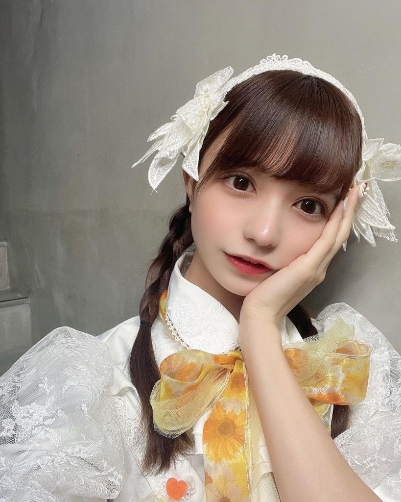 A young woman with braided hair adorned with a large white bow, wearing a frilly white dress and a floral-patterned scarf, poses with her hand on her cheek against a neutral background.