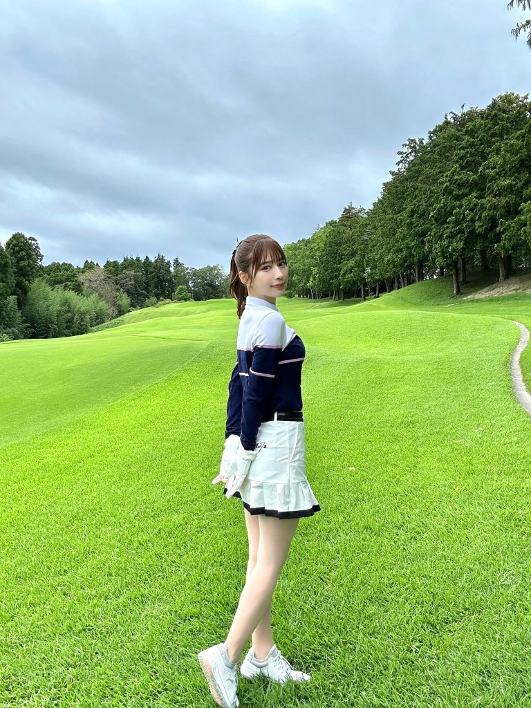 A woman stands on a lush green golf course, wearing a sporty outfit with a navy blue top and a white skirt. She has her back turned slightly towards the camera, showcasing the landscape of trees and a cloudy sky in the background.