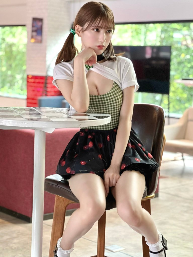 A young woman with styled hair sitting on a stool, wearing a checkered top and a cherry-patterned skirt, appears contemplative in a well-lit indoor space.
