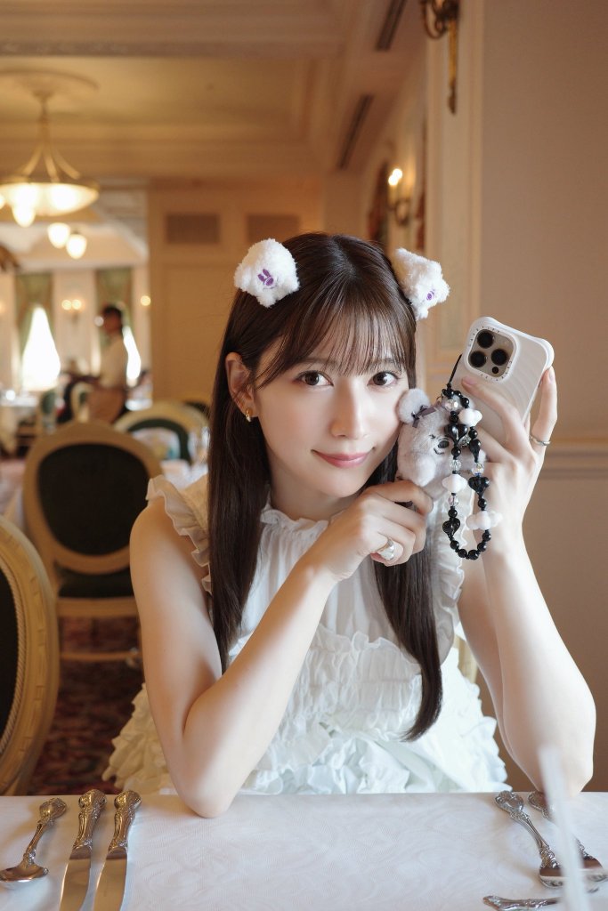 A young woman wearing a white ruffled dress and cute animal ears poses while holding her phone and a small toy at a dining table in an elegant restaurant.