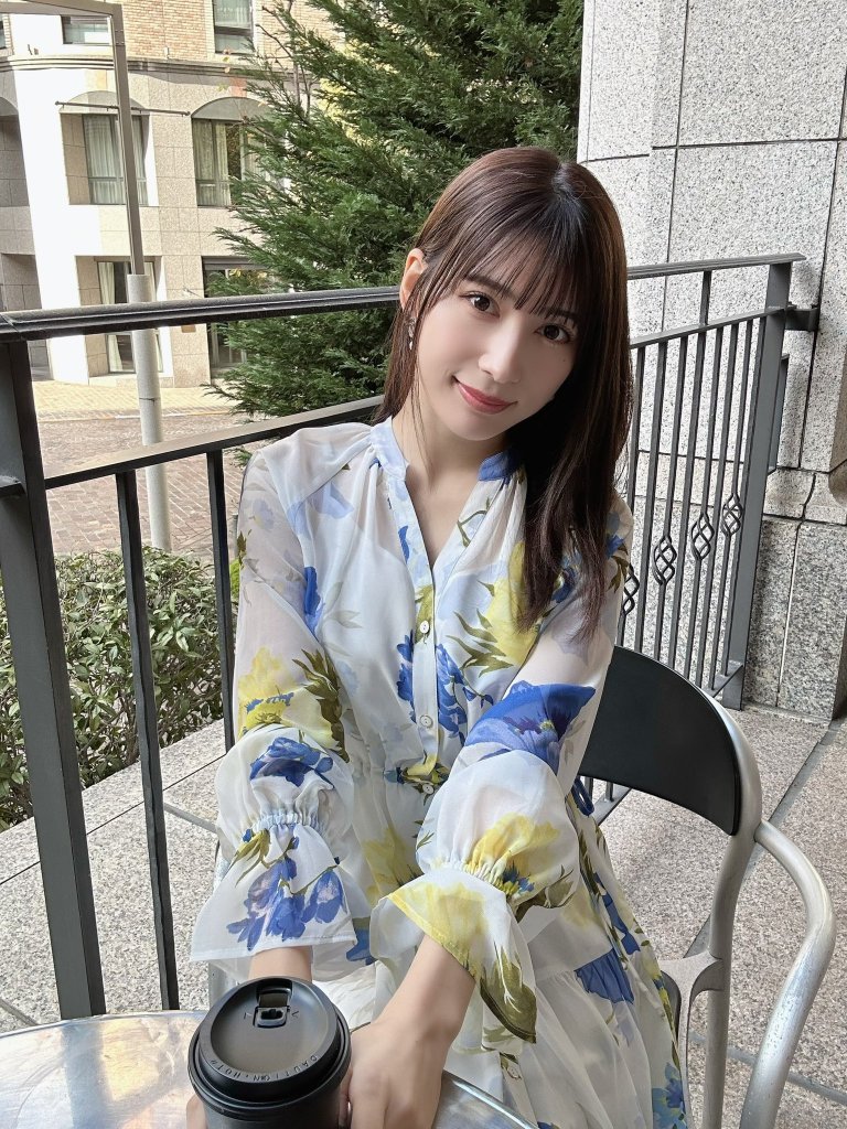 A person sitting at a table wearing a floral dress, holding a black coffee cup, with greenery and modern buildings in the background.