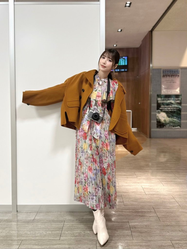 A woman wearing a colorful floral dress, a brown coat, and white boots, posing in a well-lit indoor space.