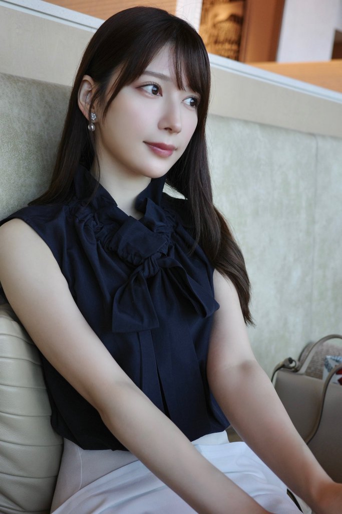A young woman with long, dark hair, wearing a black sleeveless blouse with a bow tie, sitting on a beige couch with a neutral background.